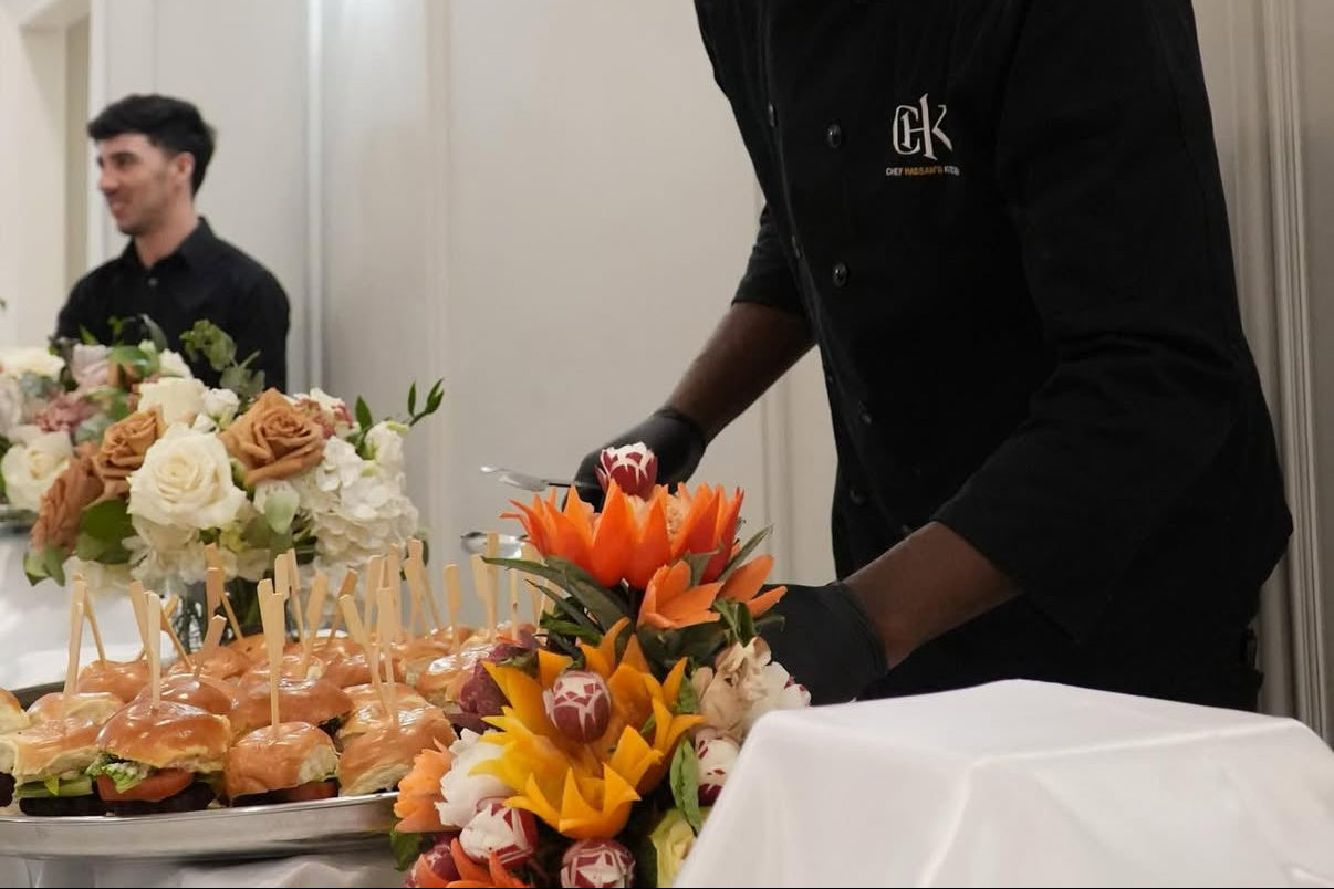 Man in a black chef's coat arranging flowers at a table with food.