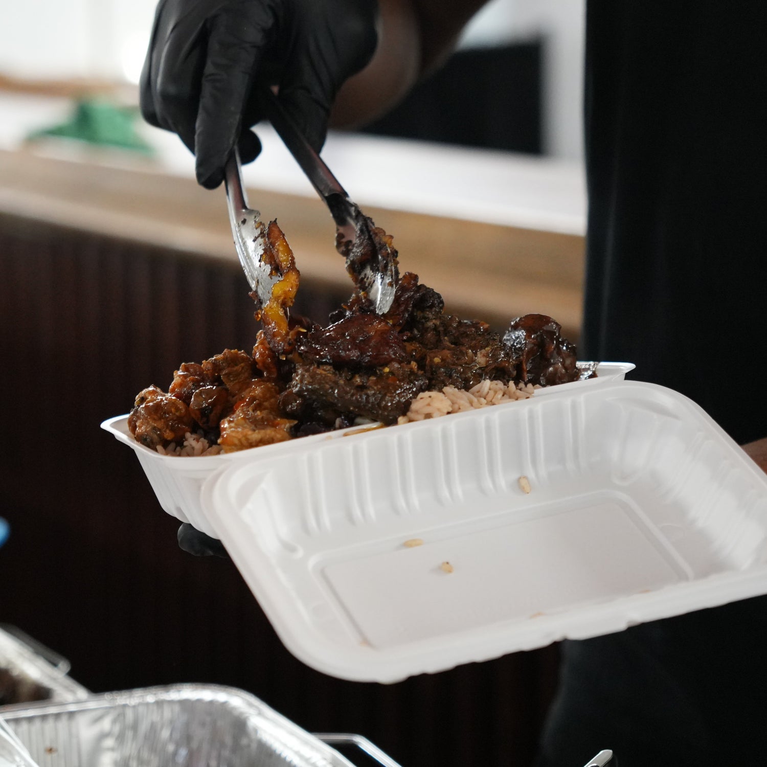Person serving food from a container with tongs, wearing black gloves.