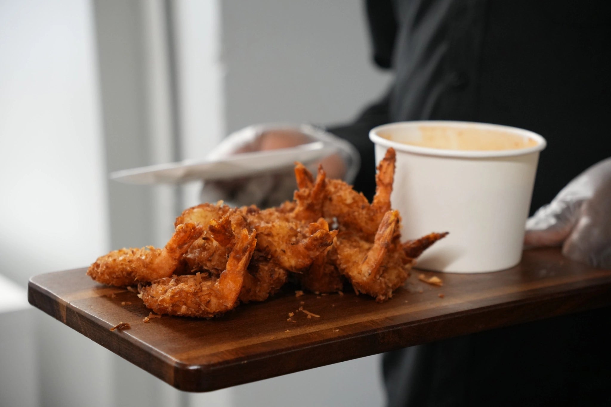 Fried coconut shrimp on a wooden board with a dipping sauce, held by a person in a blurred background.